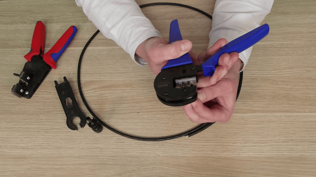 Close-up of hands using a specialised MC4 solar connector crimping tool to prepare a cable. Other solar installation tools are visible in the background.